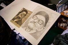 Kamerūnietis dailininkas, belaukiant prieglobsčio suteikimo, piešia portretus iš laikraščio. Kamerūnietis dailininkas, belaukiant prieglobsčio suteikimo, piešia portretus iš laikraščio.