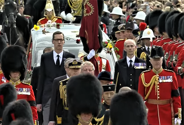 Aukštas vyras juodu fraku ir medalių juosta kadro dešinėje - ištikimasis karalienės pažas Paulas Whybrew.
